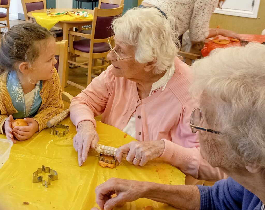 An elderly woman and a young girl sit at a table with baking supplies, making treats and exchanging stories of friendship, while another elderly woman looks on.