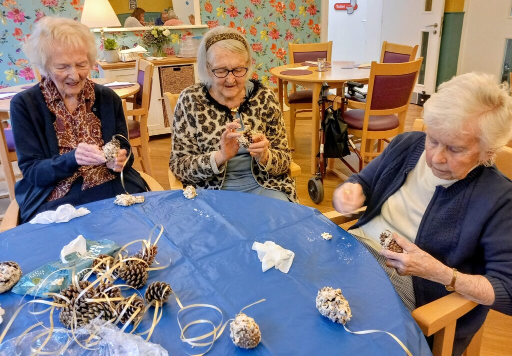 Three elderly women enjoy crafting together, coating pinecones with a white substance in a brightly decorated room. Their friendship is clear as they share laughter and creativity.