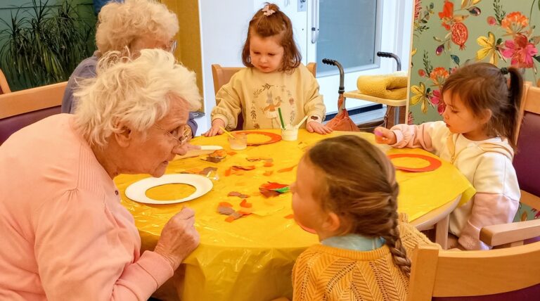 Elderly women and young children sit gathered around a table draped with a yellow cloth, crafting together with paper and paint, their friendship evident in every creative moment.