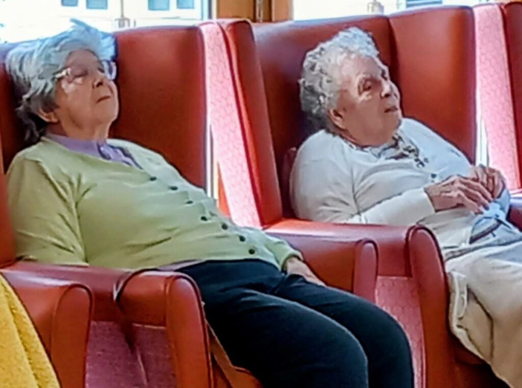 Two elderly women with grey hair sit and relax in red armchairs, possibly reminiscing or gently dozing, as soft music drifts through the brightly lit room.