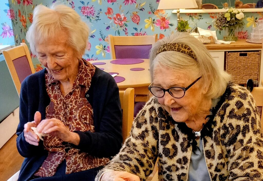 Two elderly women sit at a table crafting together, smiling and shaping materials with their hands, in a brightly decorated room that reflects the warmth of their friendship.
