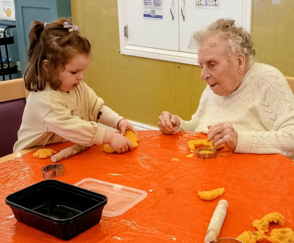 A young girl and an elderly woman sit at a table covered in orange plastic, making shapes from yellow dough with biscuit cutters and rolling pins, sharing laughter and companionship.