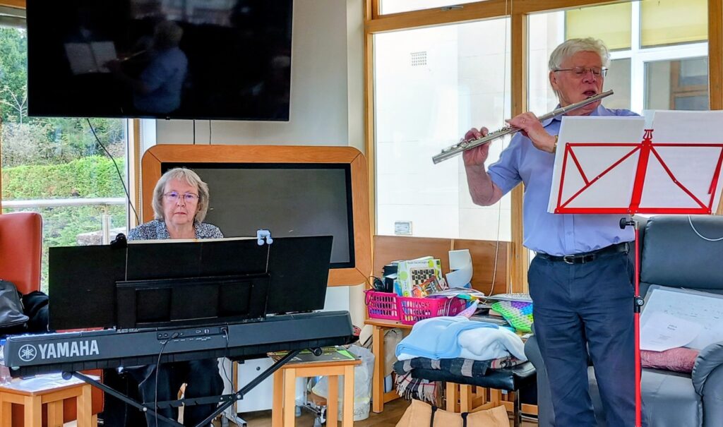 An elderly woman plays a Yamaha keyboard while an elderly man stands beside her playing the flute; both read sheet music in a sunlit room as melodies fill the space with memories.
