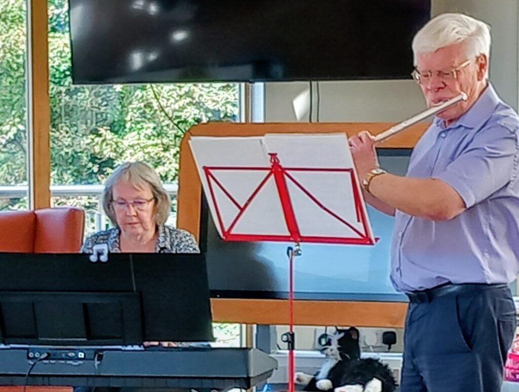 A man plays a hornpipe on the flute while a woman plays an electric keyboard. Their sheet music rests on a music stand, as a stuffed cat sits nearby, adding charm to their shared musical memories.