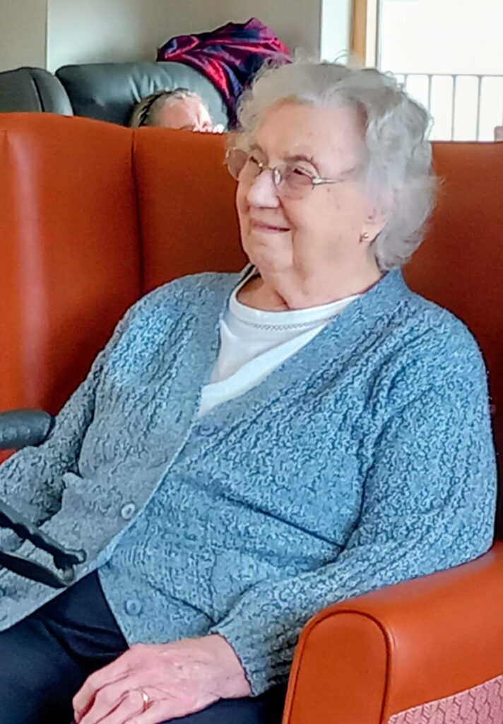 An elderly woman with grey hair and spectacles sits in an orange armchair, dressed in a blue cardigan, smiling gently as if reminiscing about music.