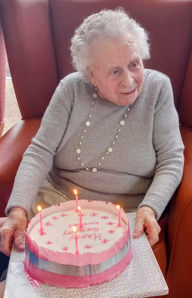 Elderly woman seated in an armchair, holding a birthday cake with lit candles and 'Happy Birthday' written on it.
