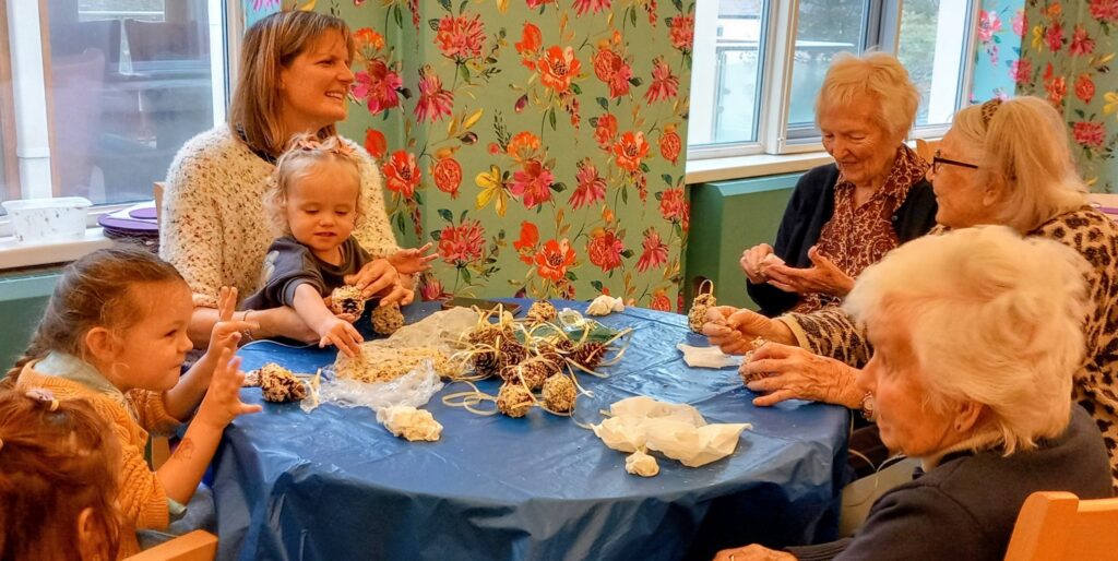 Elderly women and young children sit around a table covered with a blue cloth, making crafts together and sharing companionship in a brightly decorated room.