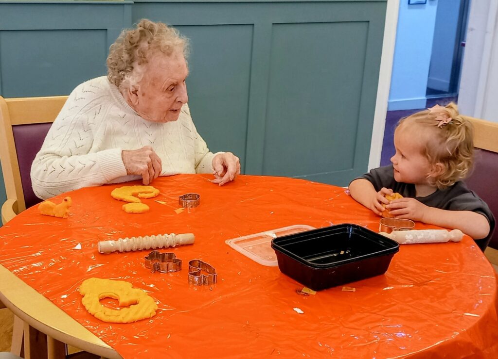 An elderly woman and a young girl sit at a table covered with orange dough and biscuit cutters, enjoying a crafting activity that celebrates their warm friendship.