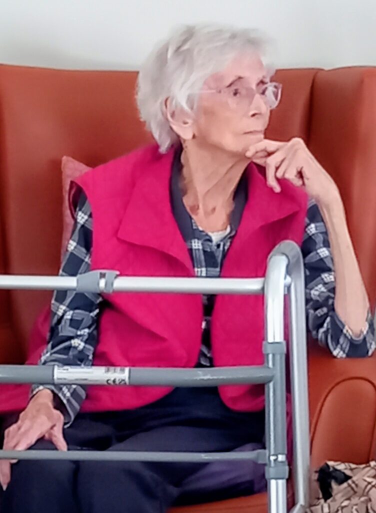 Elderly woman with white hair, wearing spectacles, a pink waistcoat, and a checked shirt sits on an orange chair next to a walker, resting her chin on her hand as if lost in thought.