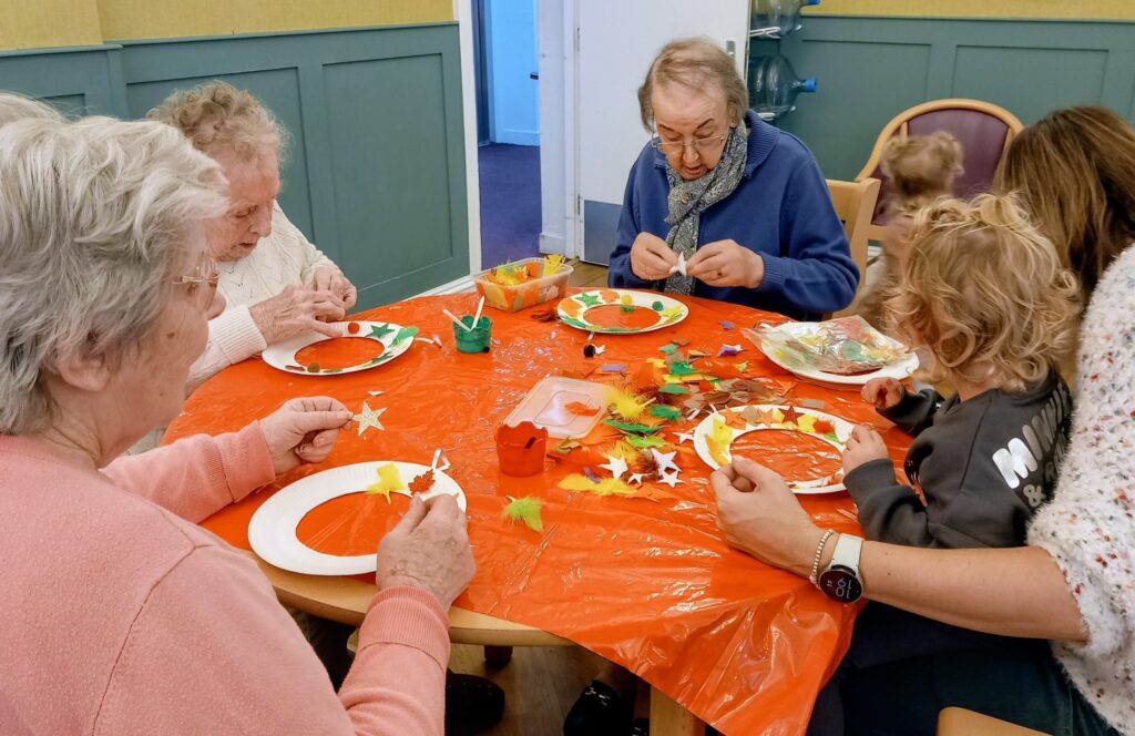 Elderly people and young children sit around a table covered with craft materials, making decorations together in a brightly lit room, their friendship evident in every creative moment.