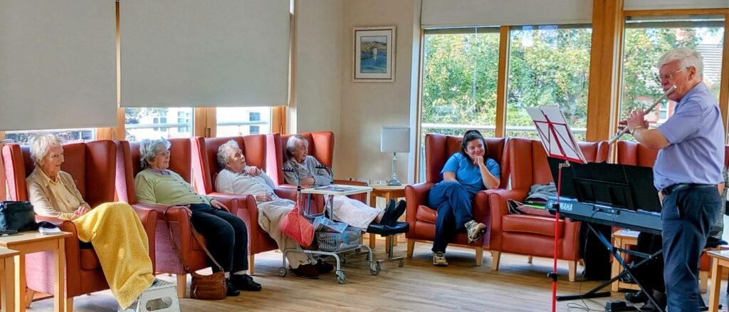 An elderly man plays the flute beside a keyboard, filling the bright communal room with melodies and evoking memories for four elderly women and a woman in scrubs seated on armchairs.