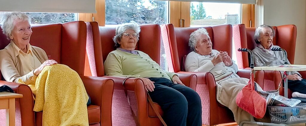 Four elderly women sit in high-backed chairs by a window, reminiscing in a brightly lit room, as the distant sound of music brings warmth to their gathering. A walking frame and basket are nearby.