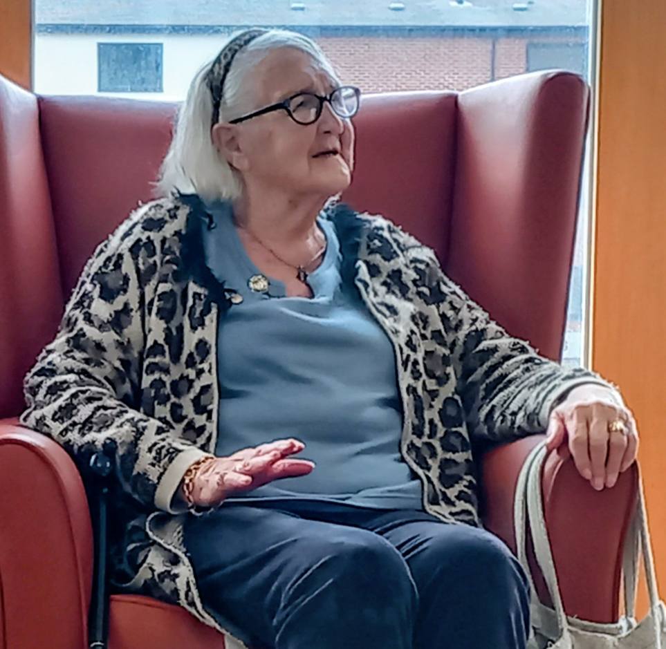 Elderly woman with white hair and glasses sits in a high-backed red chair at Lear House, wearing a leopard print cardigan over a blue top, with a handbag resting on her left arm.