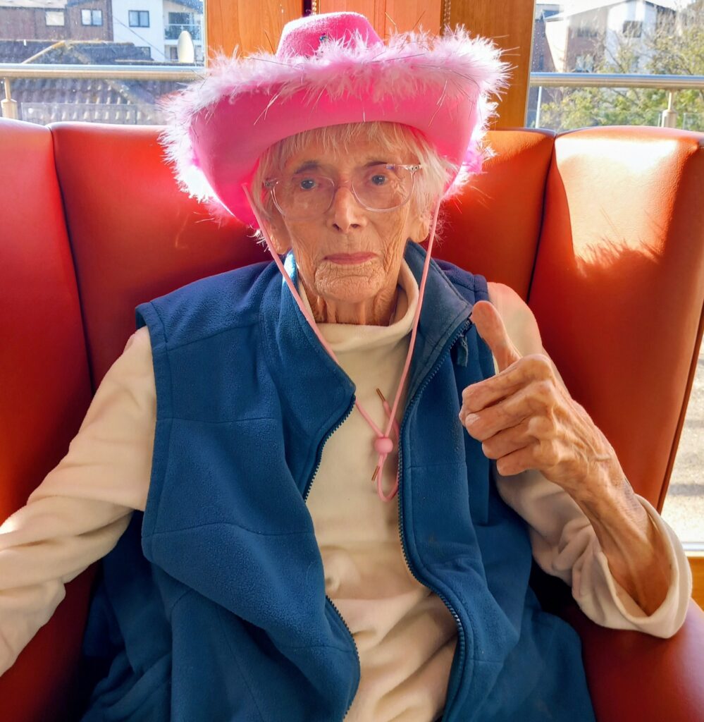 Elderly person with glasses wearing a pink cowboy hat and blue waistcoat, seated on an orange chair at Lear House, giving a thumbs up during a Cowboy Afternoon of music and fun.