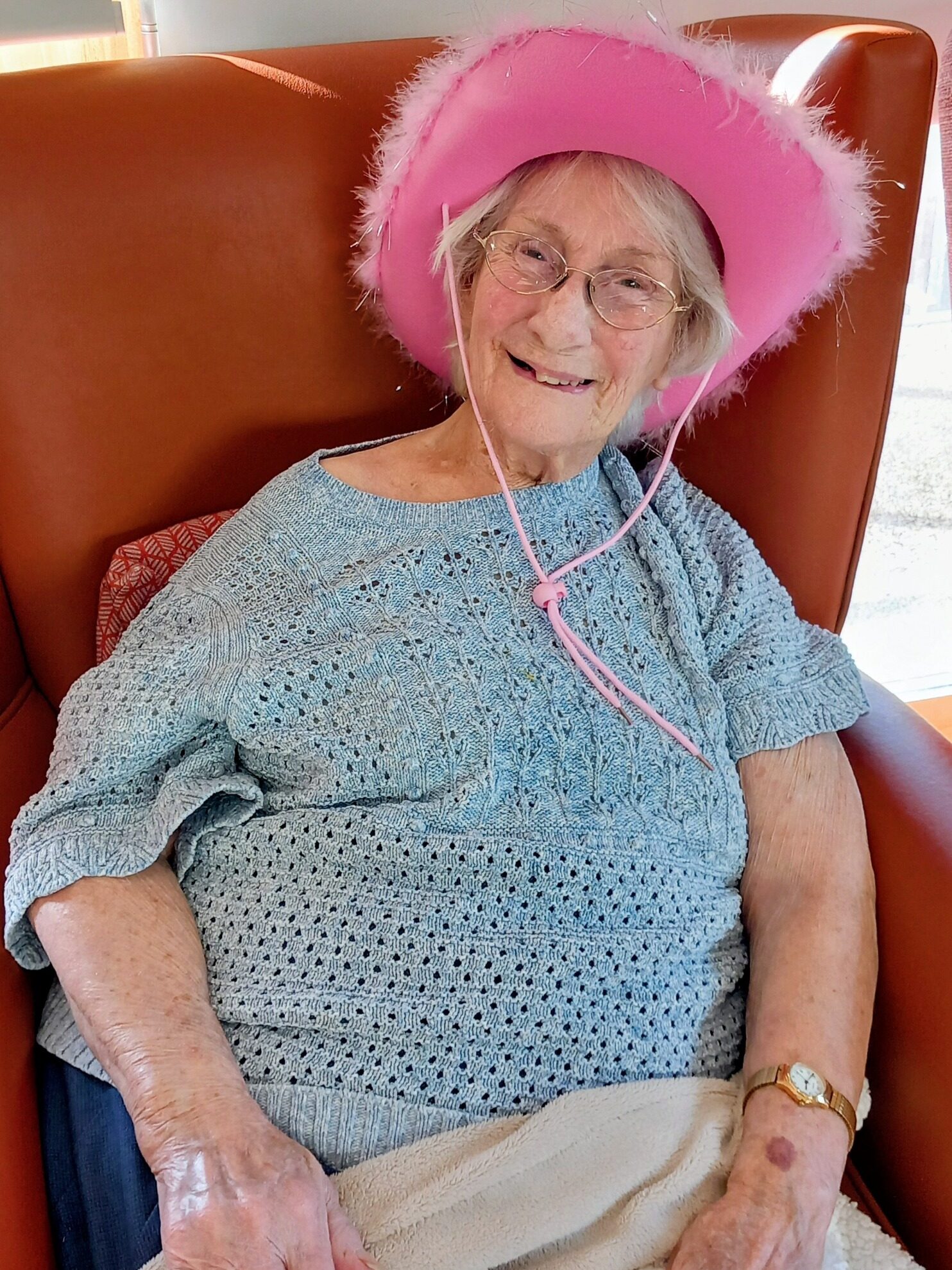 An older woman wearing spectacles and a pink cowboy hat sits in a brown chair at Lear House, smiling and covered with a light blanket—ready to enjoy a Cowboy Afternoon.