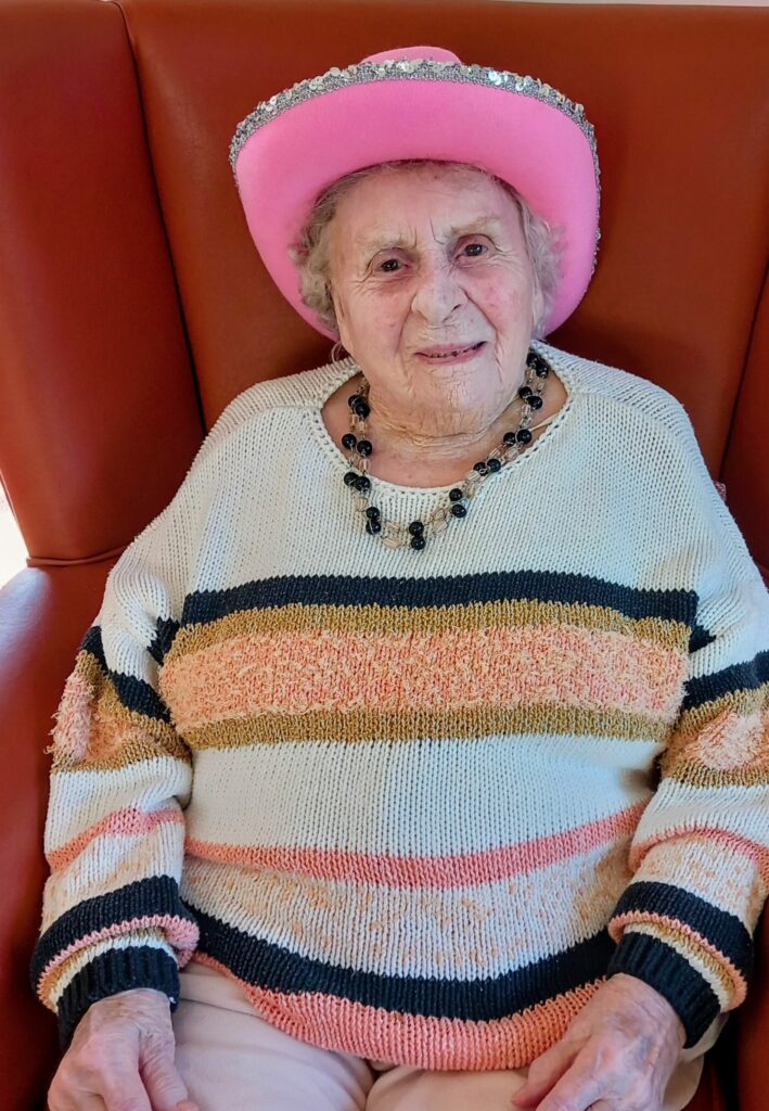 Elderly woman wearing a pink hat with silver trim, striped jumper, and beaded necklace, seated in a brown chair at Lear House during Cowboy Afternoon.