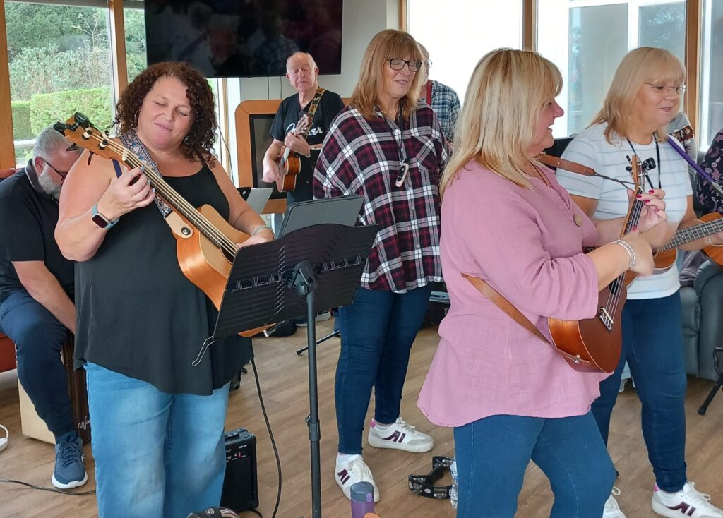 Six members of the ukulele band 64 N’Ukes are playing stringed instruments indoors; five are standing while one is seated, with music stands and a television visible in the background.