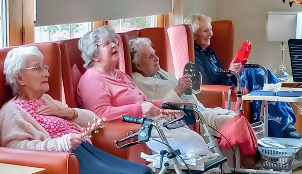Four elderly women from The 64 N’Ukes sit in armchairs at a care home, playing musical instruments and smiling together, with walking aids and personal belongings close by.