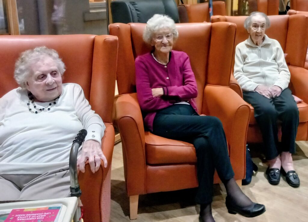 Three elderly women sit in orange armchairs in Lear House’s communal area, all wearing glasses and casual clothes, with a walking frame and a book close by—prepared for safe Bonfire Night celebrations.
