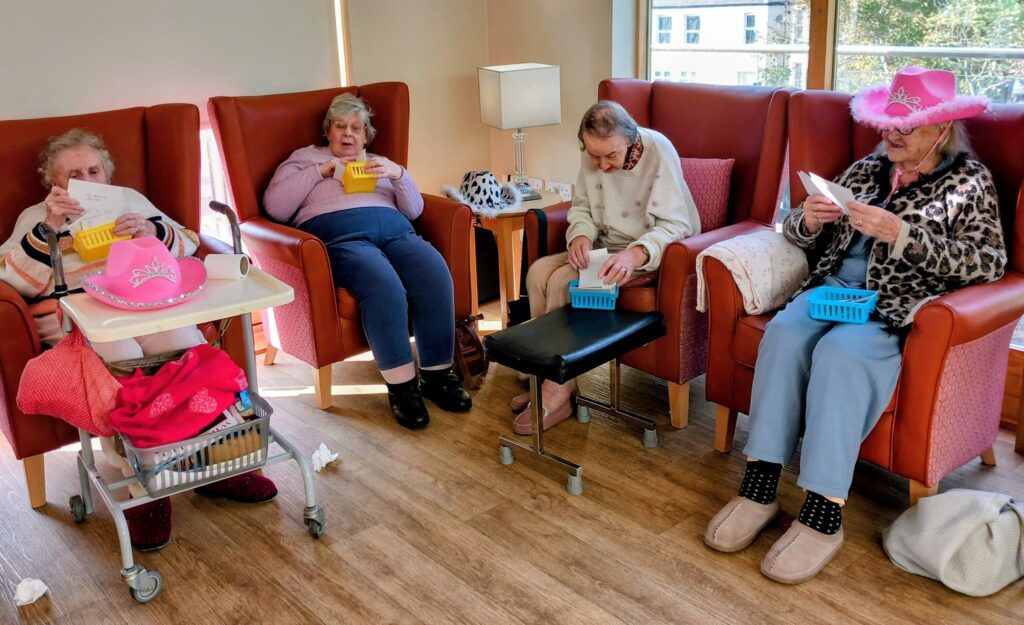 Four elderly women sit in armchairs at Lear House, playing bingo with cards and baskets in a brightly lit room. One woman sports a large pink hat, bringing extra flair to their cheerful cowboy-themed afternoon.
