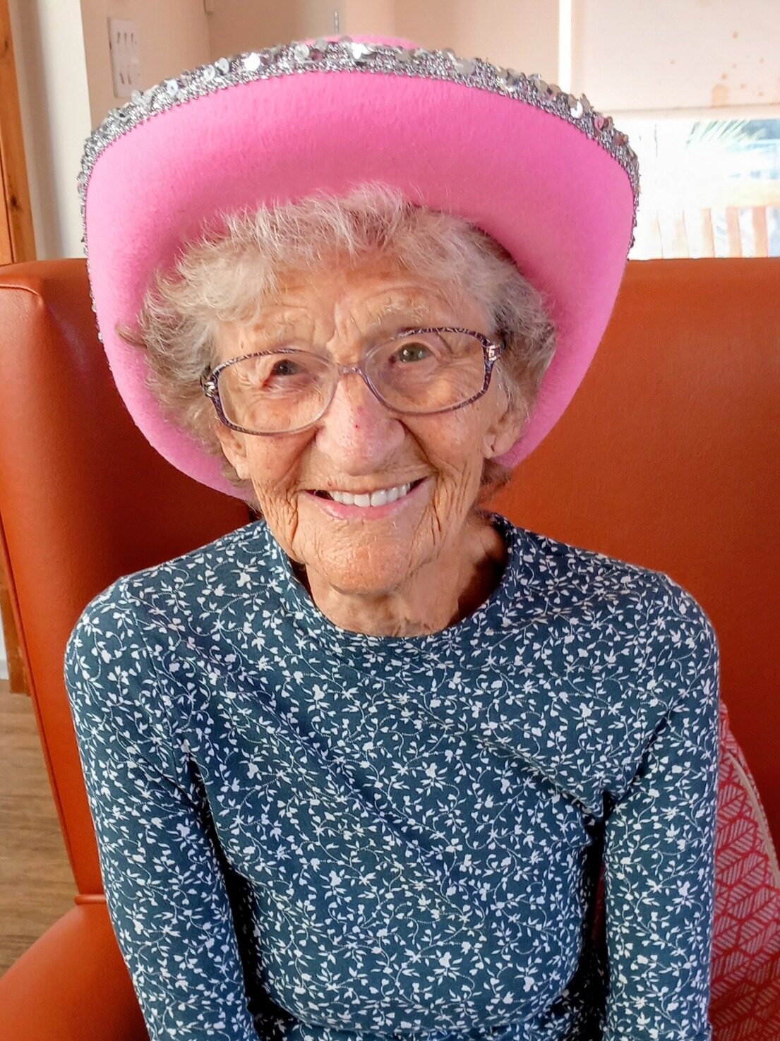Elderly woman wearing glasses and a pink cowboy hat sits in a red chair, smiling at the camera during a lively Cowboy afternoon at Lear House.