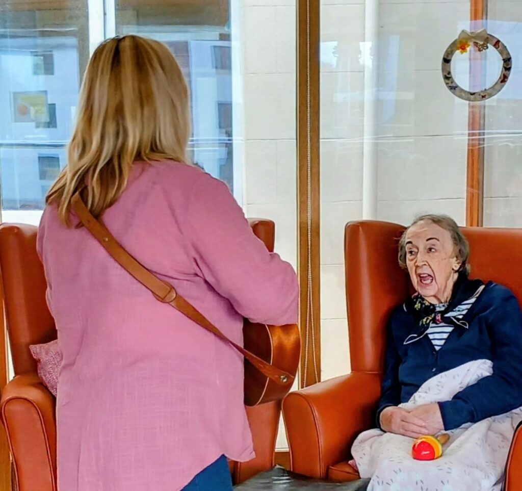 A woman with a guitar stands facing an elderly woman seated in an armchair, who seems to be singing or speaking, as if joining in with the strumming of the 64 N’Ukes ukulele band, a blanket across her lap.
