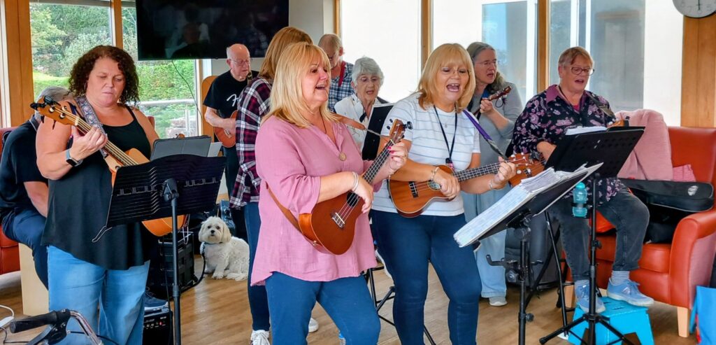 A group from 64 N’Ukes strumming ukuleles and singing together indoors, music stands in front of them, while a small white dog sits in the background amongst smiles.