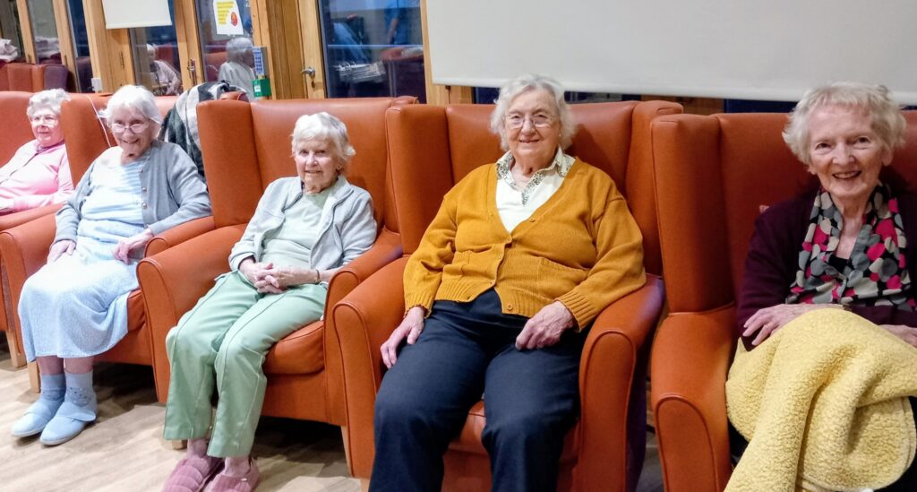 Five elderly women sit smiling in orange armchairs in a bright room at Lear House, featuring wooden floors and large windows, ready to enjoy safe celebrations this Bonfire Night.