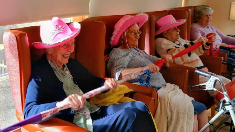 Four elderly women at Lear House sit in armchairs, wearing pink hats and holding exercise sticks, enjoying a Cowboy Afternoon group activity with lively music in a brightly lit room.
