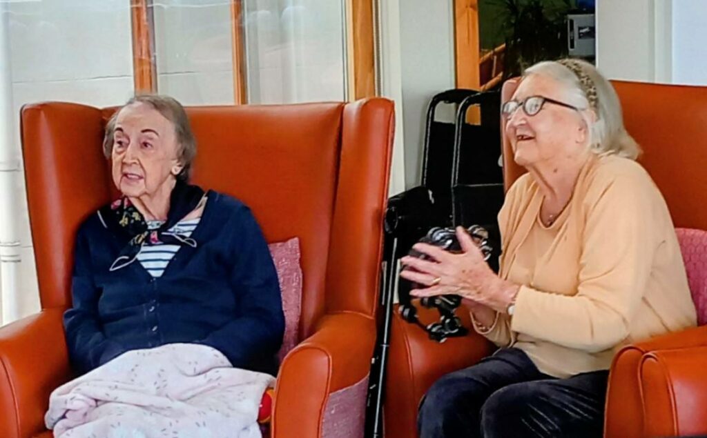 Two elderly women are seated indoors in orange chairs; one has a blanket on her lap, while the other claps and smiles, enjoying live music from 64 N'Ukes. A walking frame is visible in the background.