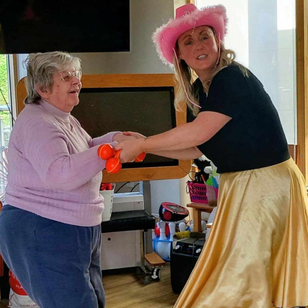 Two women dance indoors at Lear House; one sports a pink cowboy hat and yellow skirt, the other wears a pink jumper and glasses—capturing the cheerful spirit of a Cowboy Afternoon with music.
