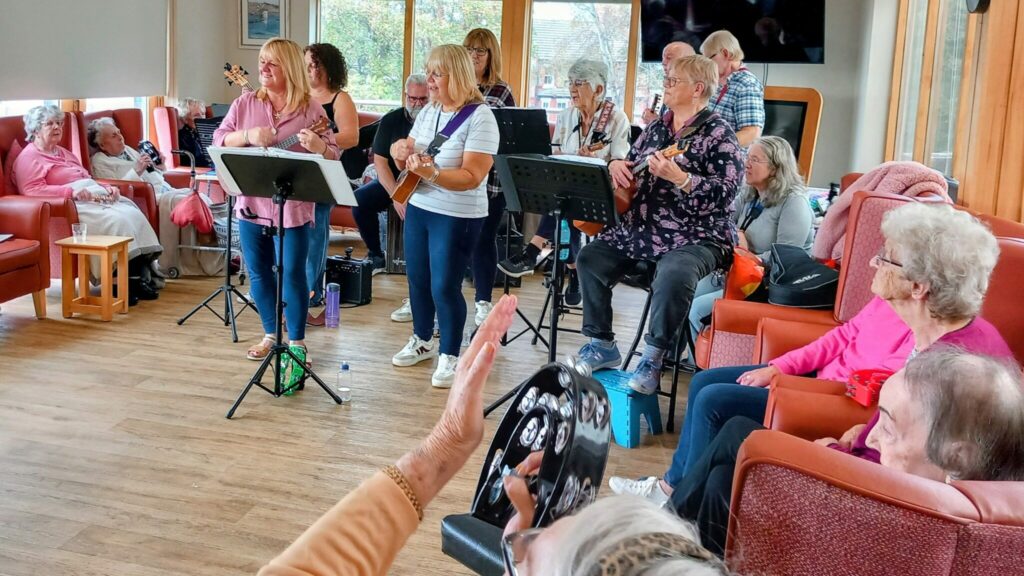 A ukulele band, 64 N’Ukes, performs for elderly people seated in a communal room; one audience member holds a tambourine and waves along to the strumming music.