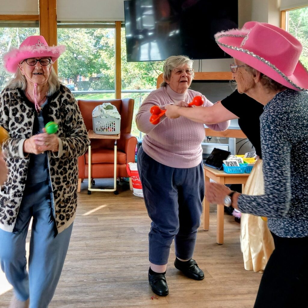 Four older women at Lear House, two wearing pink cowboy hats and shaking maracas, dance and enjoy a lively Cowboy Afternoon with music in a bright room filled with sunlight and laughter.