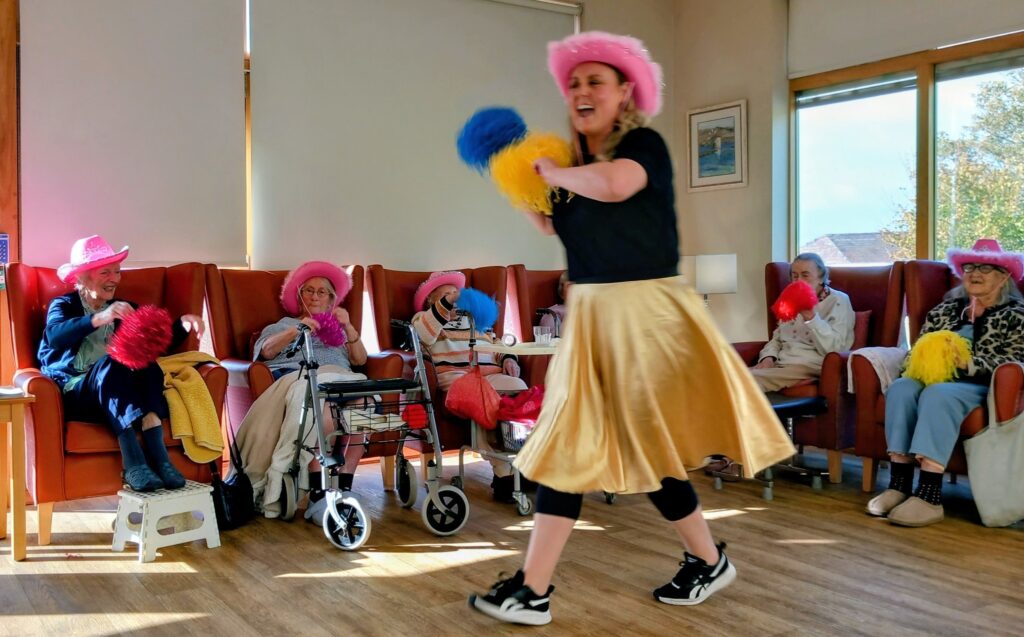 At Lear House, a woman in a gold skirt and pink hat leads elderly people, all wearing pink hats and holding pom-poms, in a cheerleading activity set to lively music in a brightly lit room.