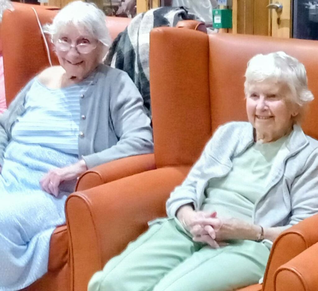 Two elderly women sit side by side in orange armchairs at Lear House, smiling—one wearing a blue dress with a grey cardigan, the other in green with a grey jacket—enjoying safe Bonfire Night celebrations.