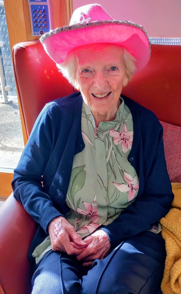 An older woman sits in a red chair at Lear House, smiling. She wears a pink cowboy hat, a floral blouse, and a dark cardigan as sunlight streams in—a perfect Cowboy Afternoon.