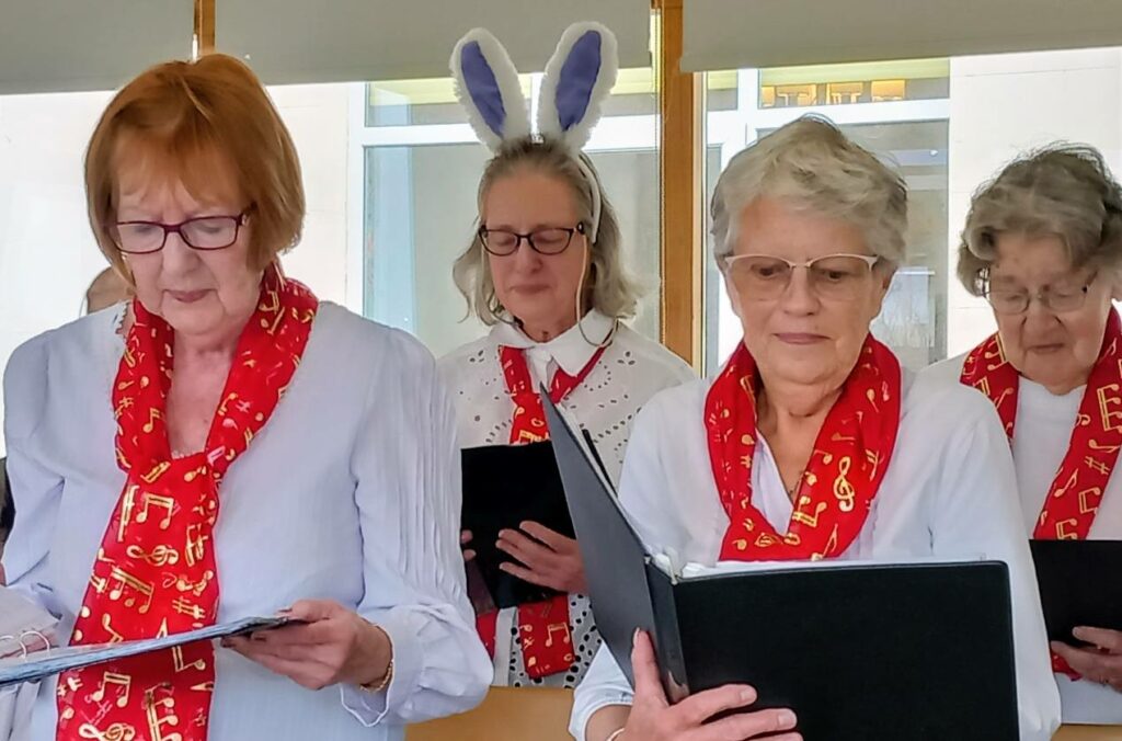 Three women from the Melrose Singers, wearing red scarves with musical notes, read from folders; the woman in the middle wears bunny ears. Another member is partly visible in the background at Lear House.