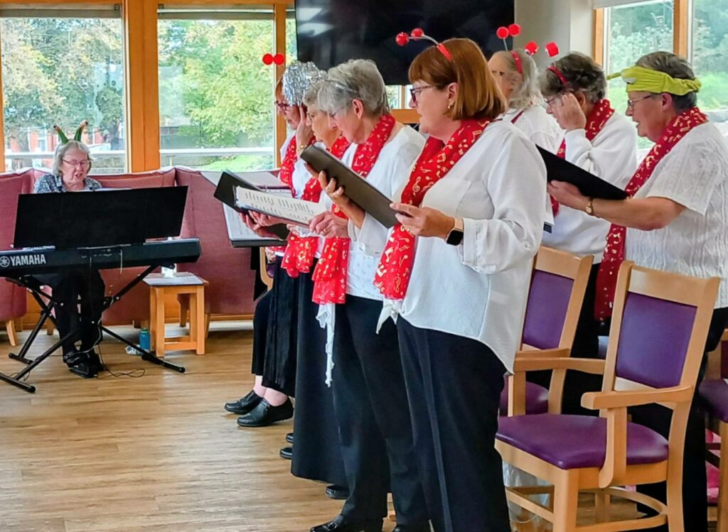 The Melrose Singers, a group of older women in red scarves and white blouses, sing from songbooks at Lear House, accompanied by a pianist on a Yamaha keyboard in a bright room with large windows.