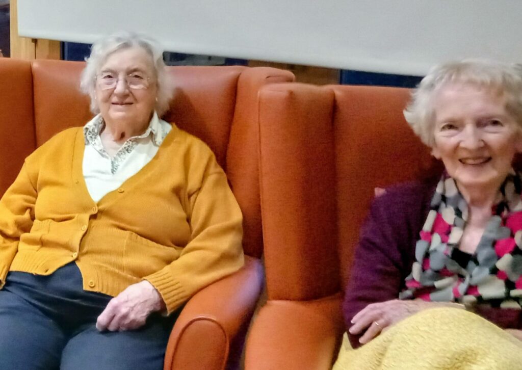 Two elderly women sit side by side on orange armchairs at Lear House, both smiling at the camera. One wears a yellow cardigan; the other has a patterned scarf and a cream blanket, enjoying safe celebrations this Bonfire Night.