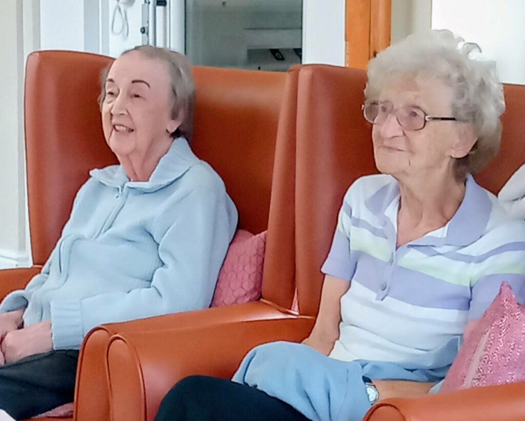 Two elderly women sit side by side in orange chairs at Lear House, facing forward. One wears a pale blue jacket, while the other, a Melrose Singers member, wears glasses and a striped polo shirt.