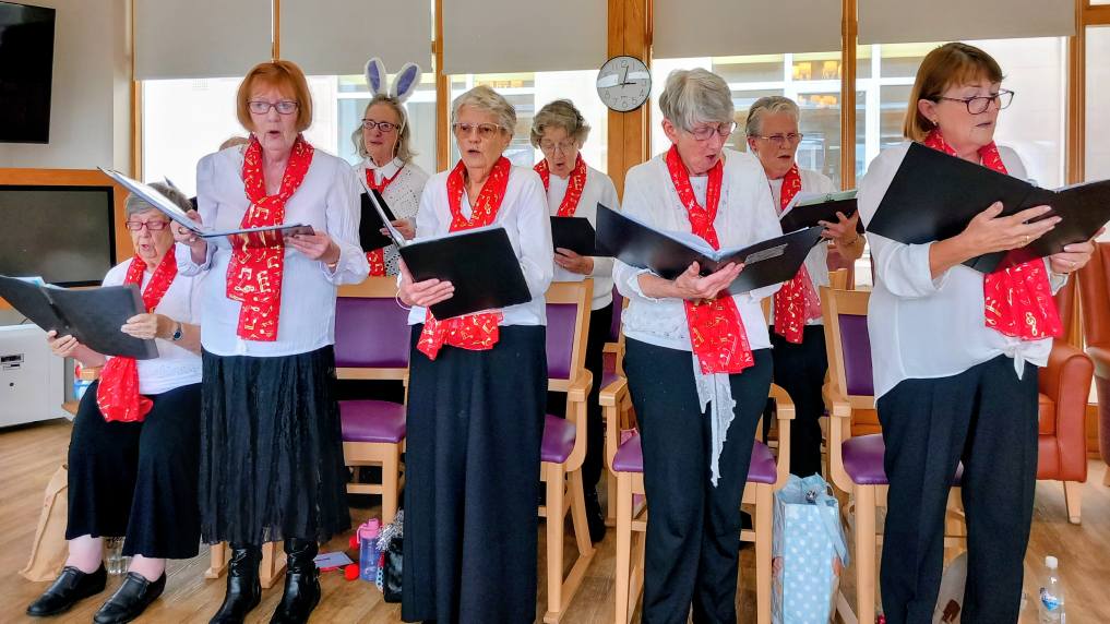 A group of older women, the Melrose Singers at Lear House, wearing red scarves and holding songbooks sing together indoors; some are standing, and one is wearing bunny ears.