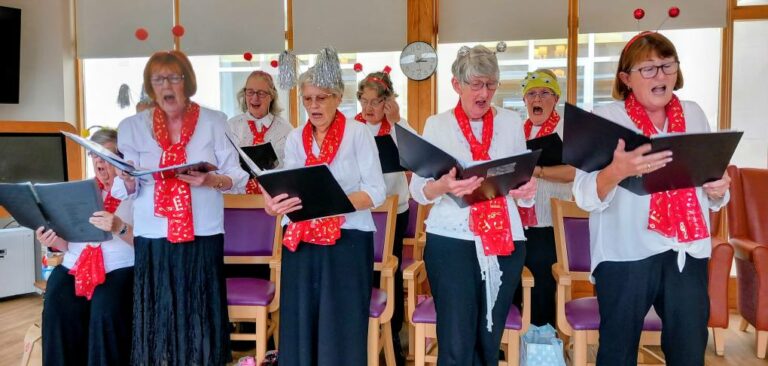 The Melrose Singers, a group of older women in red scarves and white blouses, sing together from songbooks at Lear House, gathered in a bright room with chairs and large windows.