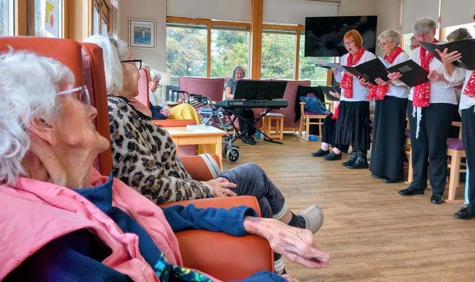 Elderly residents seated on chairs at Lear House watch the Melrose Singers perform indoors, with someone playing the keyboard in the background.