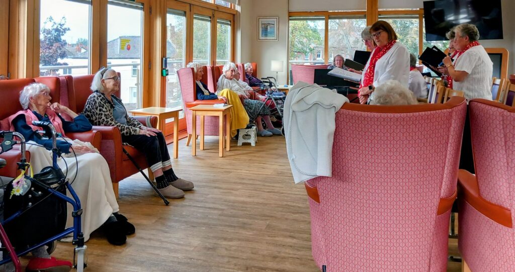 A group of elderly people sit in a lounge area at Lear House while two women from the Melrose Singers stand at the front, reading or singing from booklets.