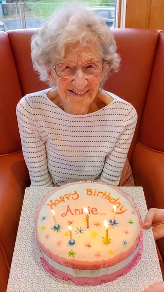 Elderly woman with white hair and glasses smiles while sitting behind a decorated birthday cake with lit candles and “Happy Birthday Annie” written on top, celebrating Annie’s special day.
