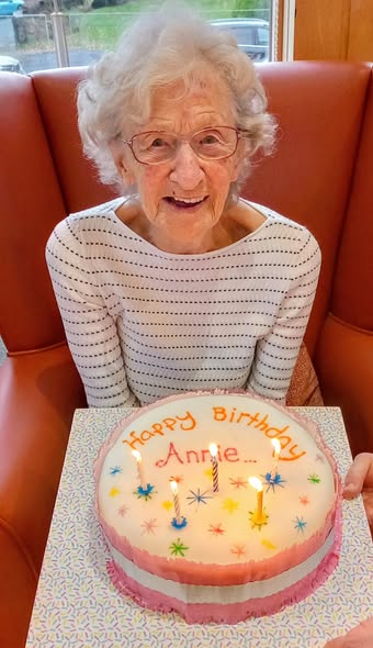 An elderly woman with grey hair and spectacles smiles as she holds a birthday cake with lit candles and "Happy Birthday Annie" written on it, celebrating with Dale.
