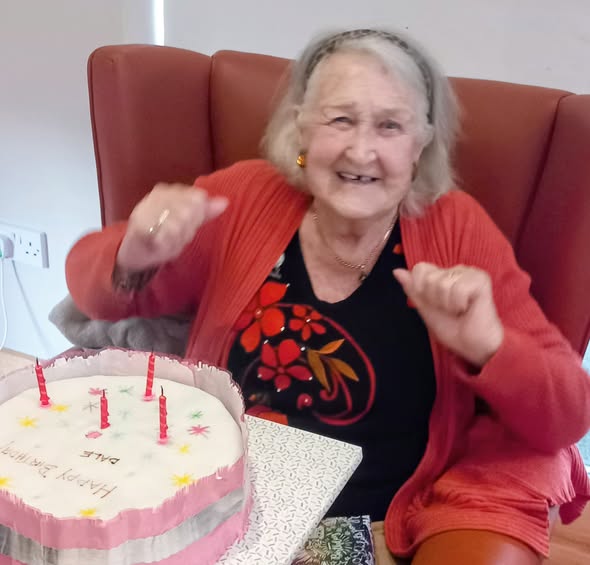 Elderly woman named Annie smiling and raising her fists in celebration while sitting next to a birthday cake with lit candles that reads "Happy Birthday".