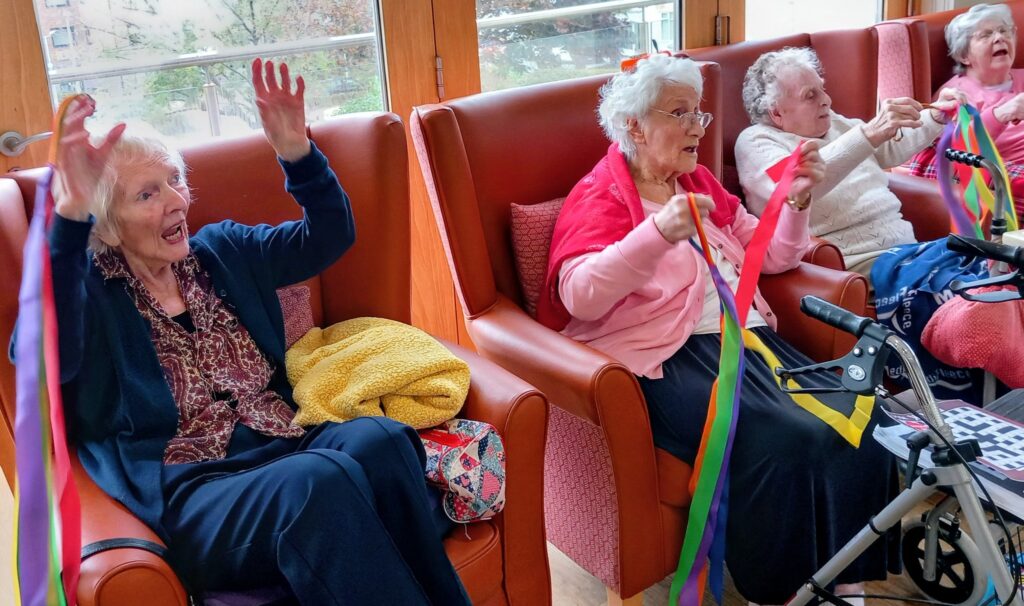 Four elderly women sit in armchairs, holding colourful ribbons and lifting their arms as part of what seems to be a group activity in a bright room.