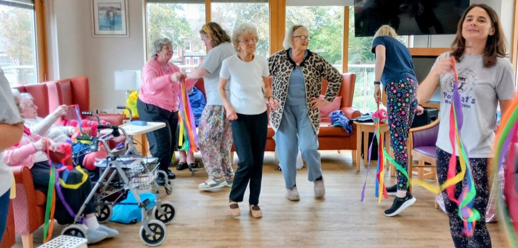 A group of elderly women and a carer take part in a ribbon dance activity in a bright, communal room with large windows and seating.