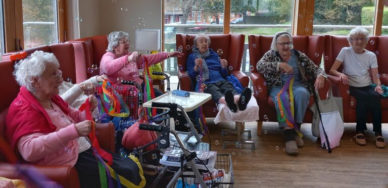 Five older women sit in armchairs in a bright room, smiling and playing with colourful ribbons and bubbles.